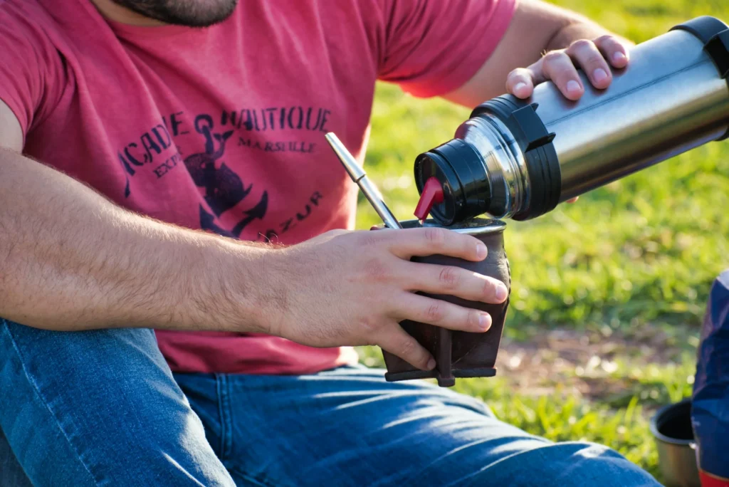 A man in a red T-shirt and jeans sitting outdoor and pouring water from a thermos into a yerba mate gourd cup with bombilla.