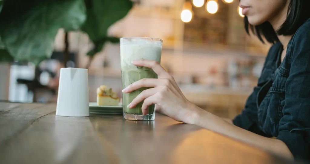 Woman sitting at a table with matcha latte, dessert, and a laptop.